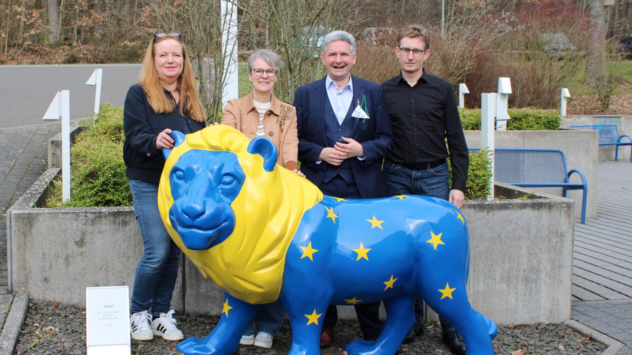 Vier Personen stehen im Freien hinter einer großen, bunt bemalten Löwenfigur. Der Löwe ist in kräftigem Blau und Gelb gestaltet und trägt gelbe Sterne auf dem Körper. Die Personen stehen dicht beieinander, einige berühren die Figur. Rechts und links befinden sich Betonpflanzkübel, dahinter sind Büsche, Wege und ein paar Sitzbänke zu sehen. Ein kleines Schild steht links neben der Figur. Geben Sie Ihr Feedback zu BizChat ab.