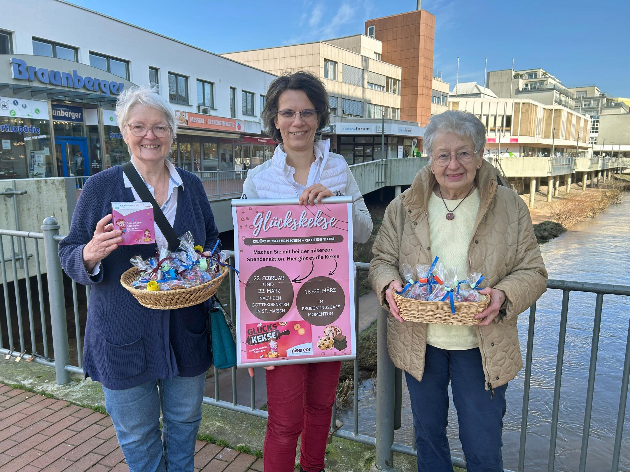 Drei Frauen stehen auf einer Brücke in der Neunkircher Innenstadt. Zwei von ihnen halten je einen Korb mit bunt verpackten Glückskeksen. Die Frau in der Mitte hält ein großes, pinkfarbenes Plakat mit Informationen zu einer Glückskeks‑Aktion. Im Hintergrund sind Geschäfte, Gebäude und die Blies zu sehen.