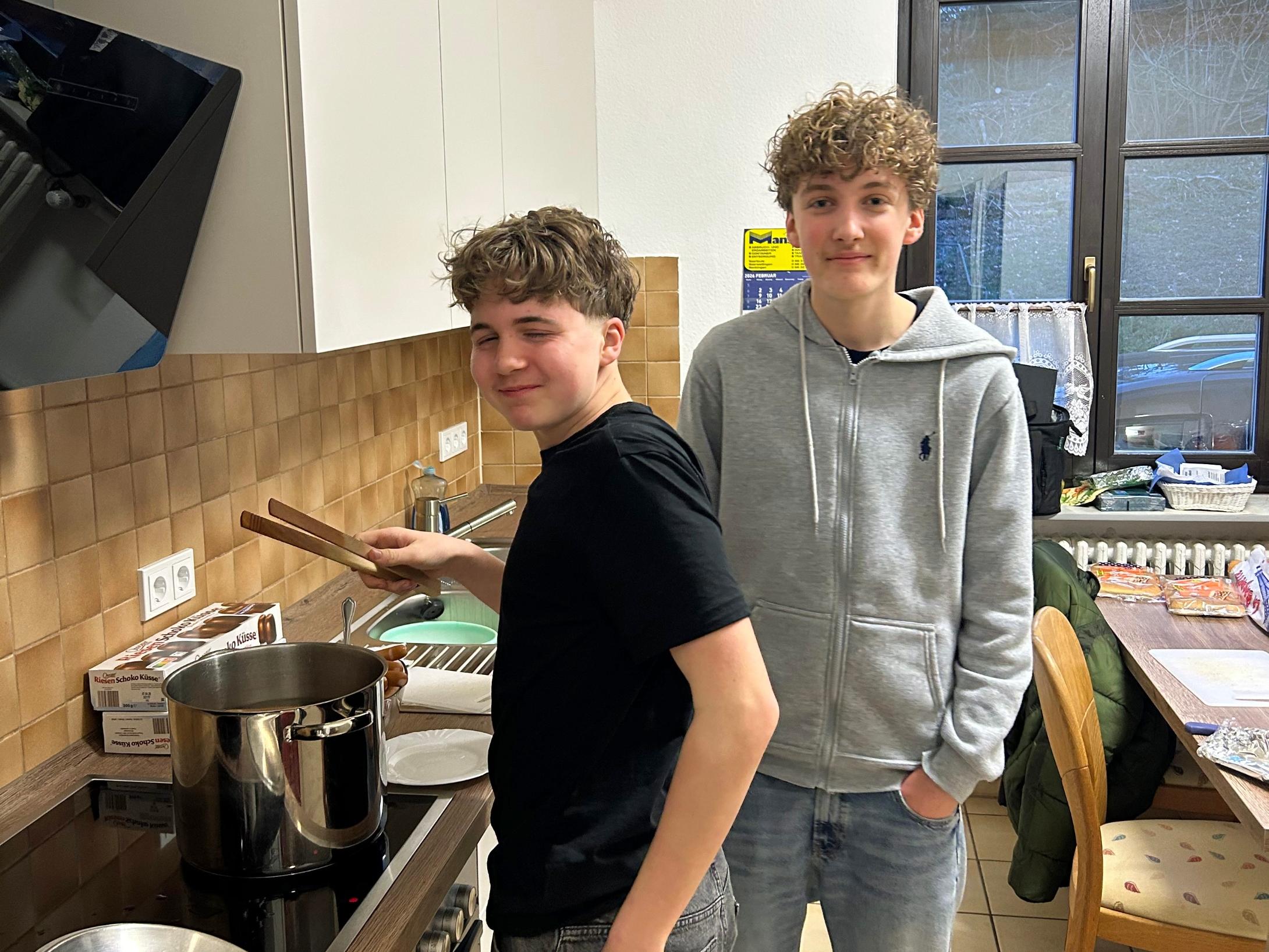 Zwei Jugendliche stehen in einer Küche. Eine Person steht am Herd und rührt mit Küchenzangen in einem großen Edelstahlkochtopf auf der Arbeitsplatte. Daneben liegen ein Topfdeckel und Zutaten wie Hotdog-Brötchen. Die zweite Person steht leicht seitlich dahinter, mit den Händen in den Hosentaschen. Im Hintergrund sind Küchenschränke, ein Fenster, ein Esstisch mit Stühlen und weitere Lebensmittel zu sehen.