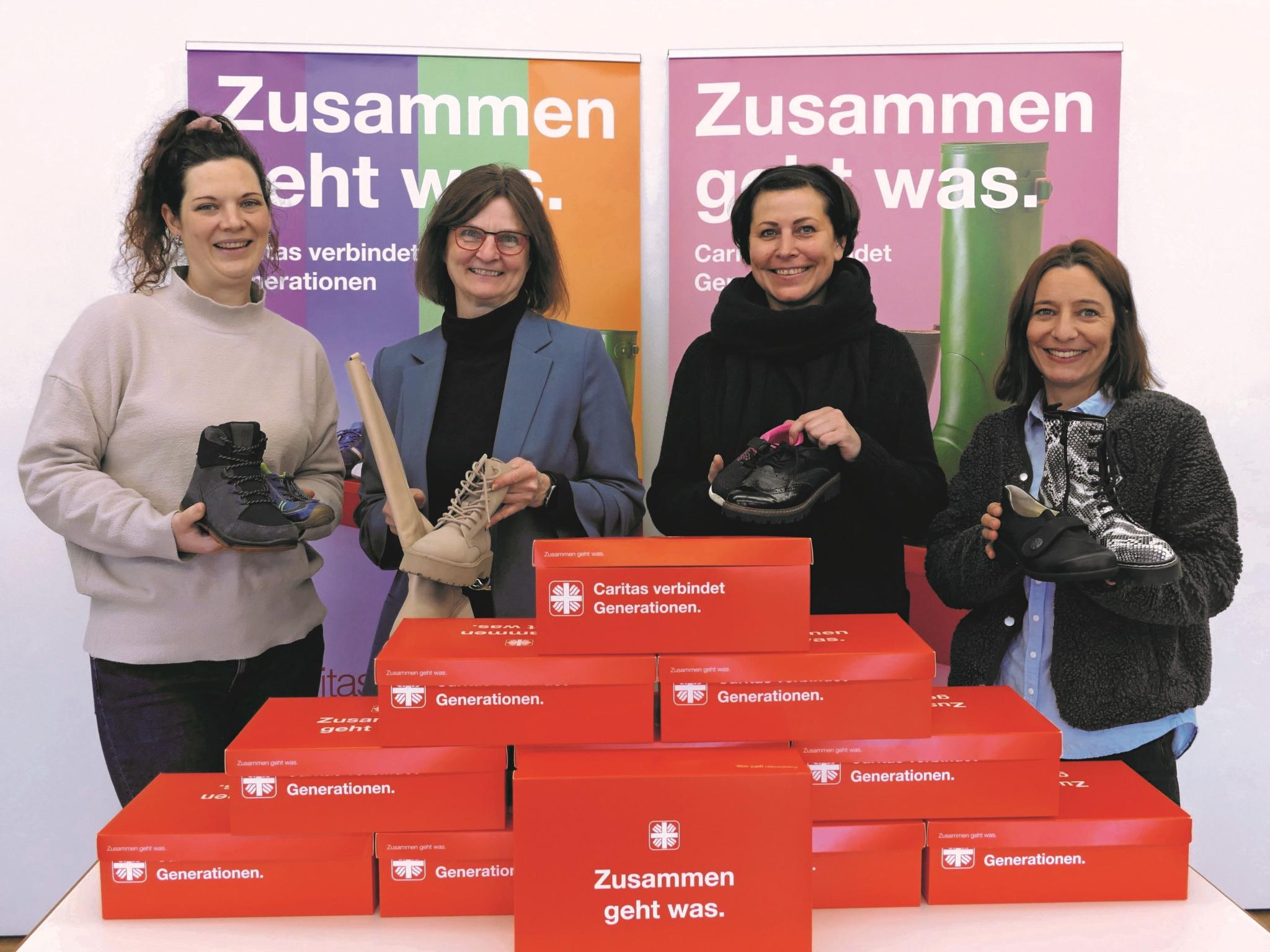 Vier Frauen lächeln in die Kamera und halten je einen Schuh hoch, vor ihnen stehen rote Schuhkartons.
