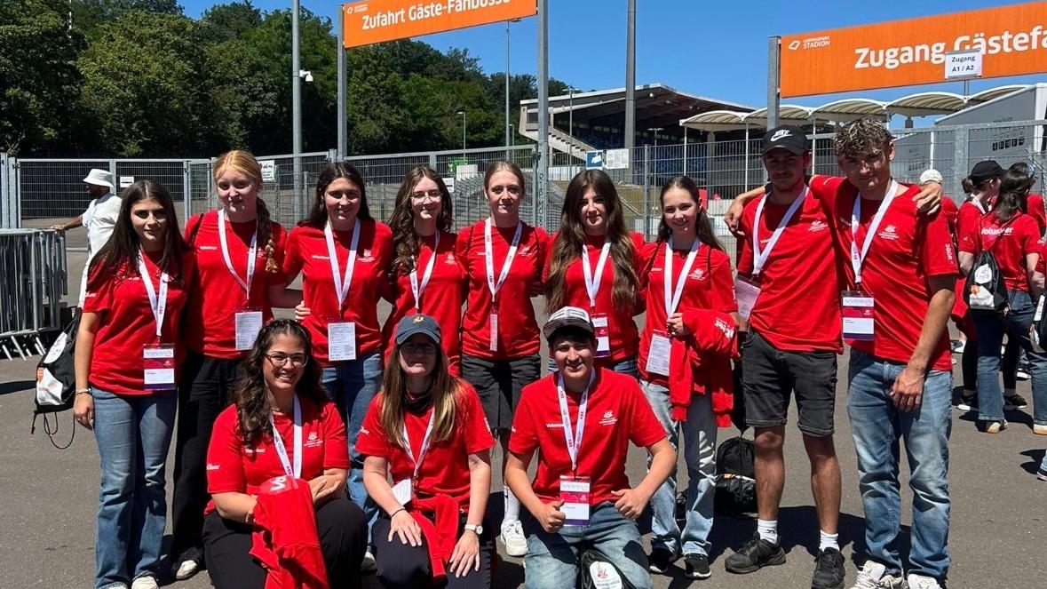 Gruppe von etwa elf Freiwilligen in roten T-Shirts mit Namensschildern und Akkreditierungen vor einem Stadioneingang. Die Personen stehen und knien für ein Gruppenfoto auf einem asphaltierten Platz. Im Hintergrund sind Zäune, Tribünen, Flutlichtmasten und orangefarbene Schilder mit den Aufschriften „Zufahrt Gäste-Fanbusse“ und „Zugang Gästefans“ zu sehen. Sonniger Tag mit klarem blauem Himmel.