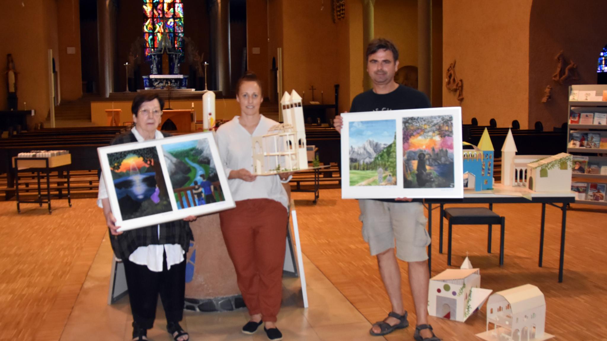 Man sieht drei Personen in einer Kirche stehen. Alle halten ein Kunstwerk der Schülerinnen und Schüler in der Hand.