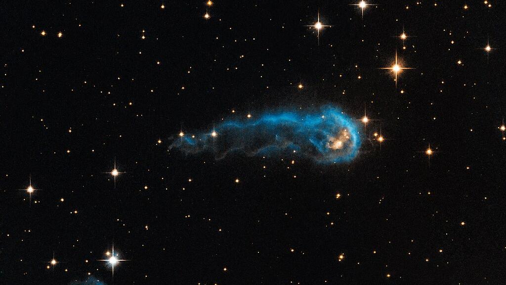 Das Weltraum-Foto zeigt das schwarze Weltall mit weißlich bis gelblich leuchtenden Sternen von etwa einem bis fünf Millimetern Größe. In der Bildmitte eine längliche, außen blau leuchtende Wolke.