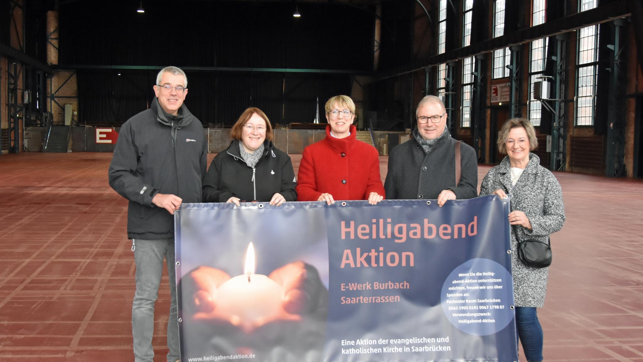 Fünf Personen stehen in einer Halle und halten das Plakat zur Heiligabend-Aktion.