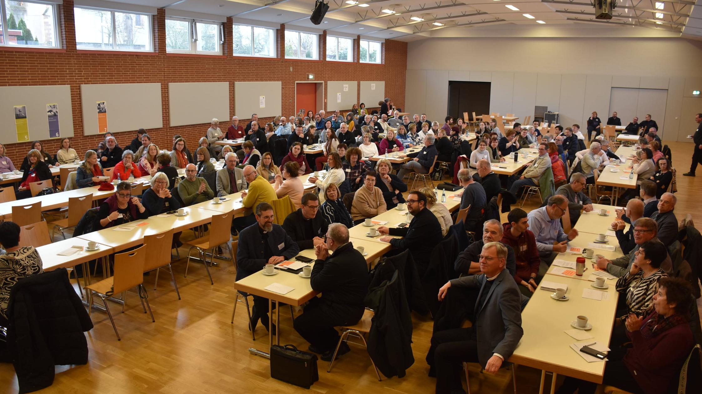 In einem großen Raum sitzen viele Menschen an langen Tischen