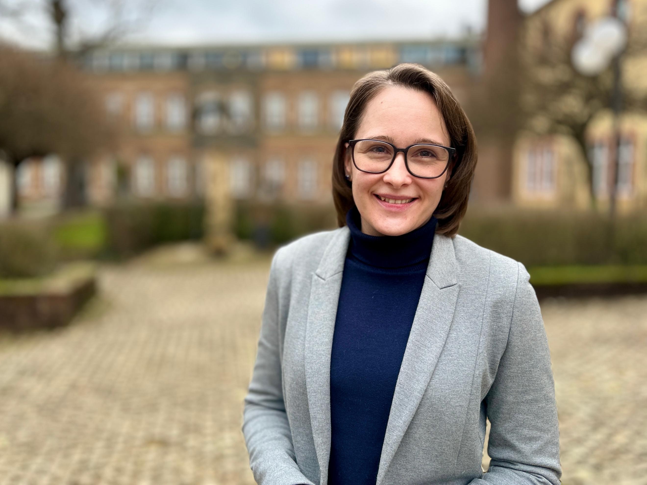 Eine Frau mit schulterlangem braunen Haar und Brille steht lächelnd im Freien vor einem historischen Backsteingebäude. Sie trägt einen grauen Blazer über einem dunkelblauen Rollkragenpullover. Der Hintergrund ist leicht unscharf, mit einem gepflasterten Weg, Hecken und Bäumen.