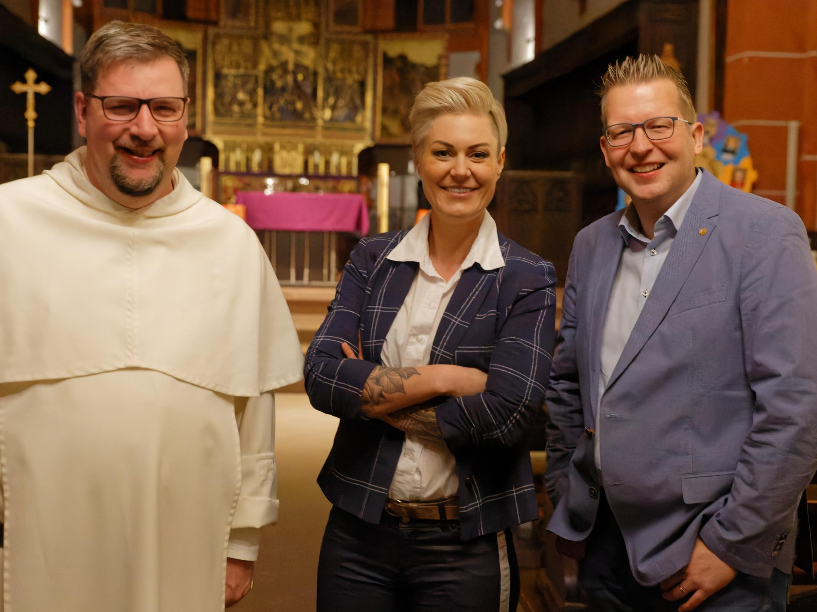 Das Foto zeigt drei Personen in einer Kirche, die freundlich in die Kamera lächeln. Von links nach rechts sind Pater Albert Seul in einer weißen Ordenskleidung, Dr. Carola Holzner aka Doc Caro in einem dunkelblauen karierten Blazer mit verschränkten Armen und Tobias Marenberg in einem hellblauen Anzug zu sehen. Der Hintergrund zeigt einen vergoldeten Hochaltar mit einer violetten Altardecke. (Alt-Text erstellt mit Unterstützung von ChatGPT)