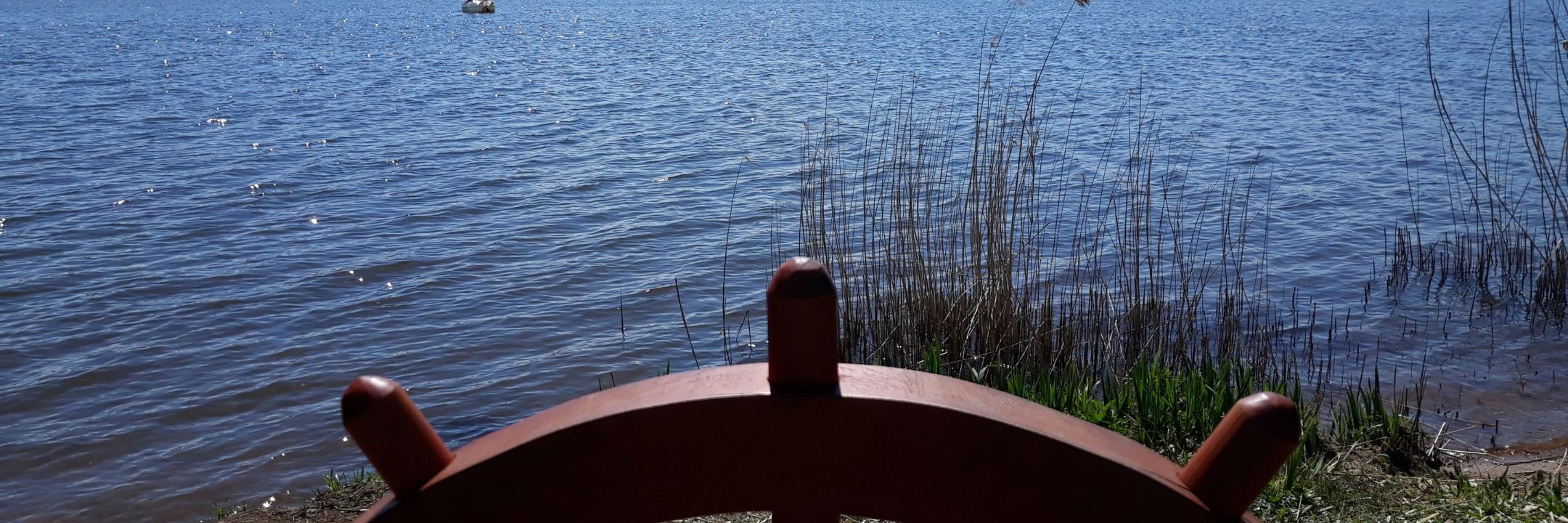 Man sieht ein eingelassenes Schiffssteuer auf einer Balustrade, dahinter der Bostalsee bei Sonnenschein