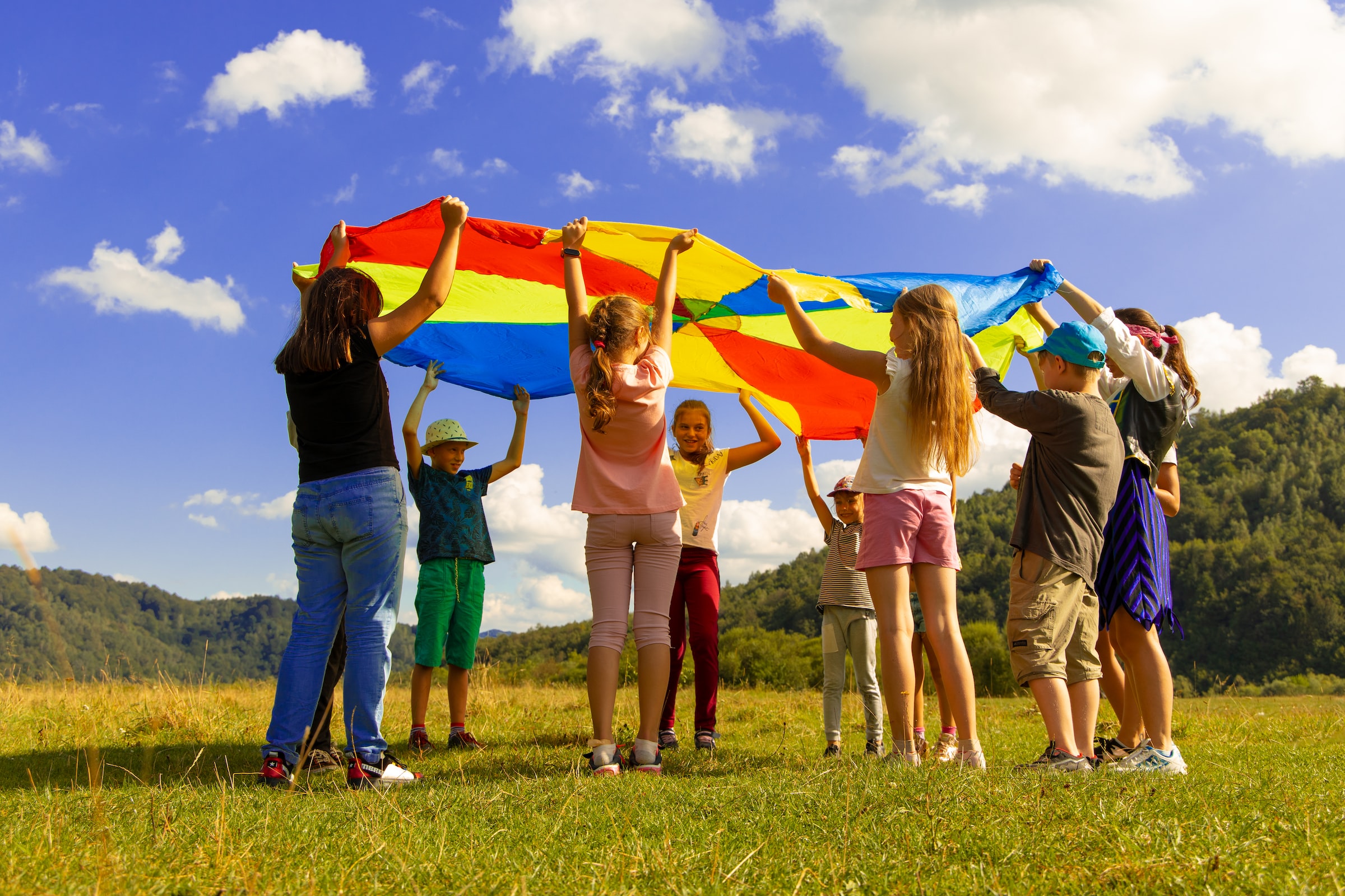 Eine Gruppe Kinder hält ein buntes 'Schwungtuch' in die Luft. Sie stehen auf einer Wiese. Der Himmel ist blau-weiß bewölkt