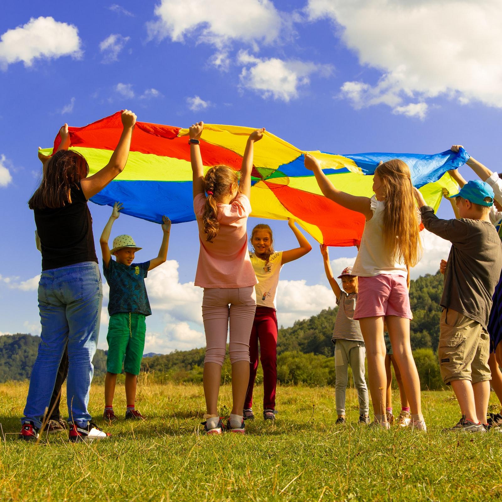 Eine Gruppe Kinder hält ein buntes 'Schwungtuch' in die Luft. Sie stehen auf einer Wiese. Der Himmel ist blau-weiß bewölkt
