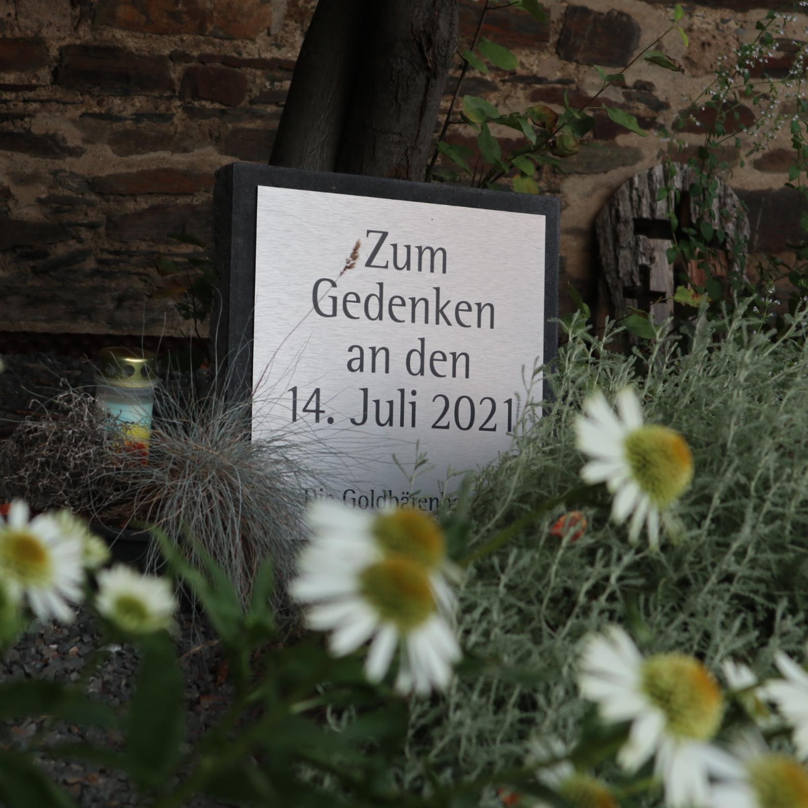 Man sieht eine Tafel auf der steht 'zum Gedenken an den 14. Juli 2021'; im Vordergrund Blumen, daneben eine Kerze.