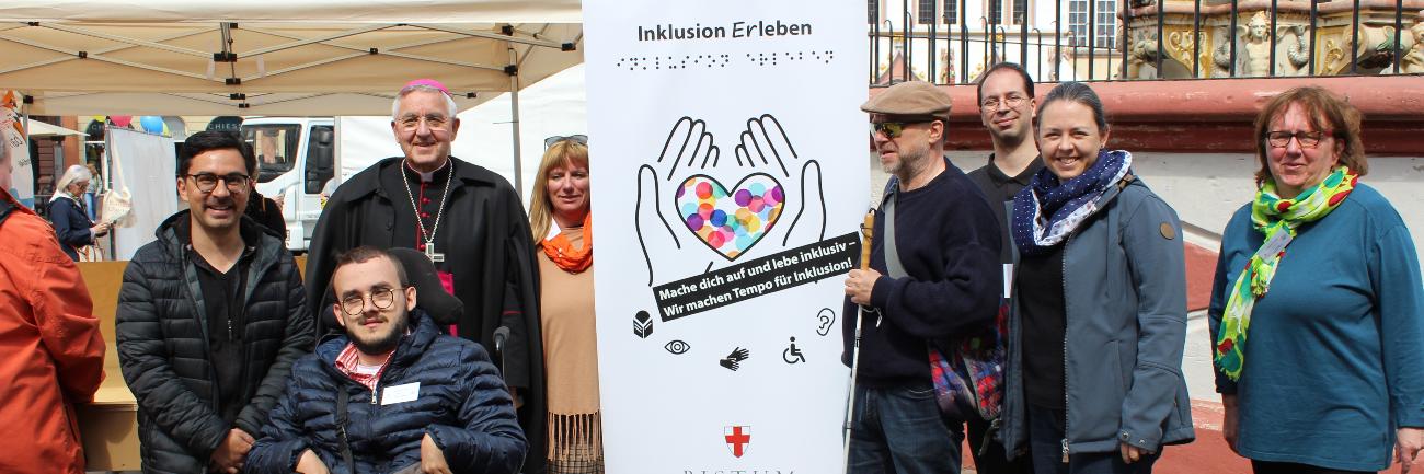 Menschen mit und ohne Behinderung stehen auf dem Hauptmarkt in Trier vor einem Aktionszelt während der Heilig-Rock-Tage 2022. In ihrer Mitte ist ein hohes Plakat zu sehen. Darauf steht oben 'Inklusion Erleben' und unten 'Bistum Trier'. Dazwischen sind die Umrisse von zwei Händen aufgedruckt, die ein Herz halten, das mit bunten Punkten gefüllt ist. Zur Gruppe gehören Weihbischof Franz-Josef Gebert, Mitarbeiterinnen und Mitarbeiter der Tonpost und aus dem Team Inklusion.