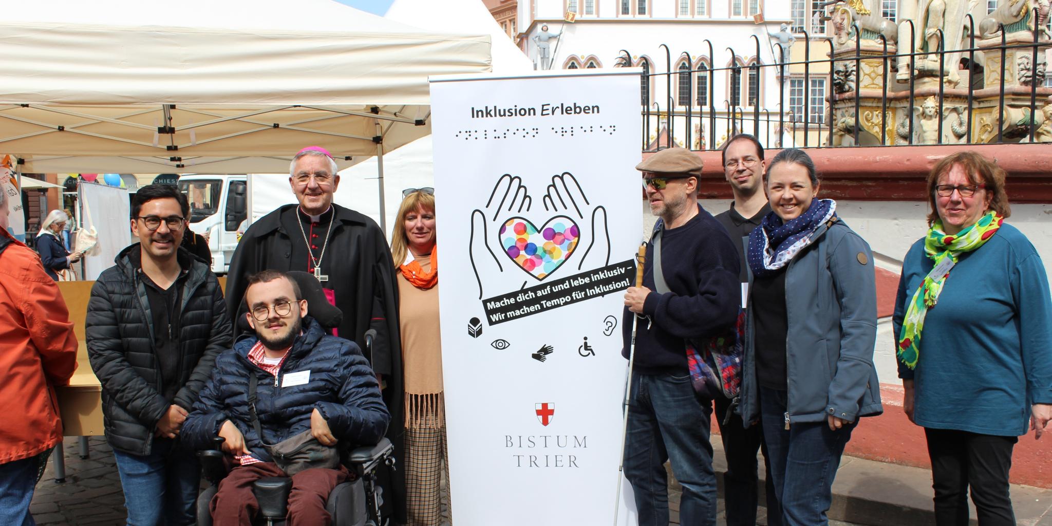 Menschen mit und ohne Behinderung stehen auf dem Hauptmarkt in Trier vor einem Aktionszelt während der Heilig-Rock-Tage 2022. In ihrer Mitte ist ein hohes Plakat zu sehen. Darauf steht oben 'Inklusion Erleben' und unten 'Bistum Trier'. Dazwischen sind die Umrisse von zwei Händen aufgedruckt, die ein Herz halten, das mit bunten Punkten gefüllt ist. Zur Gruppe gehören Weihbischof Franz-Josef Gebert, Mitarbeiterinnen und Mitarbeiter der Tonpost und aus dem Team Inklusion.