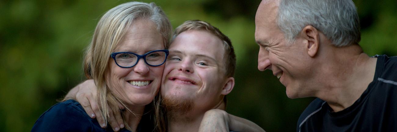 Das Bild zeigt einen Mann, eine Frau und einen jungen Mann. Alle lächeln. Die Frau und der junge Mann umarmen sich. Der junge Mann hat das Down-Syndrom.