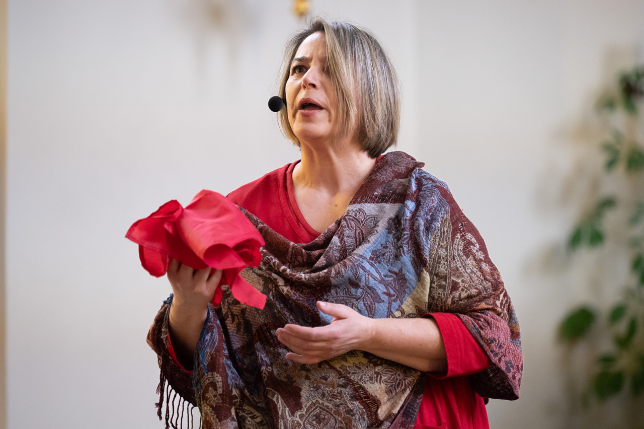 Eine Frau mit kinnlangen Haaren, rotem Kleid und einem passenden großen Schal darum spricht in ein Mikrofon, welches am Kopf befestigt ist