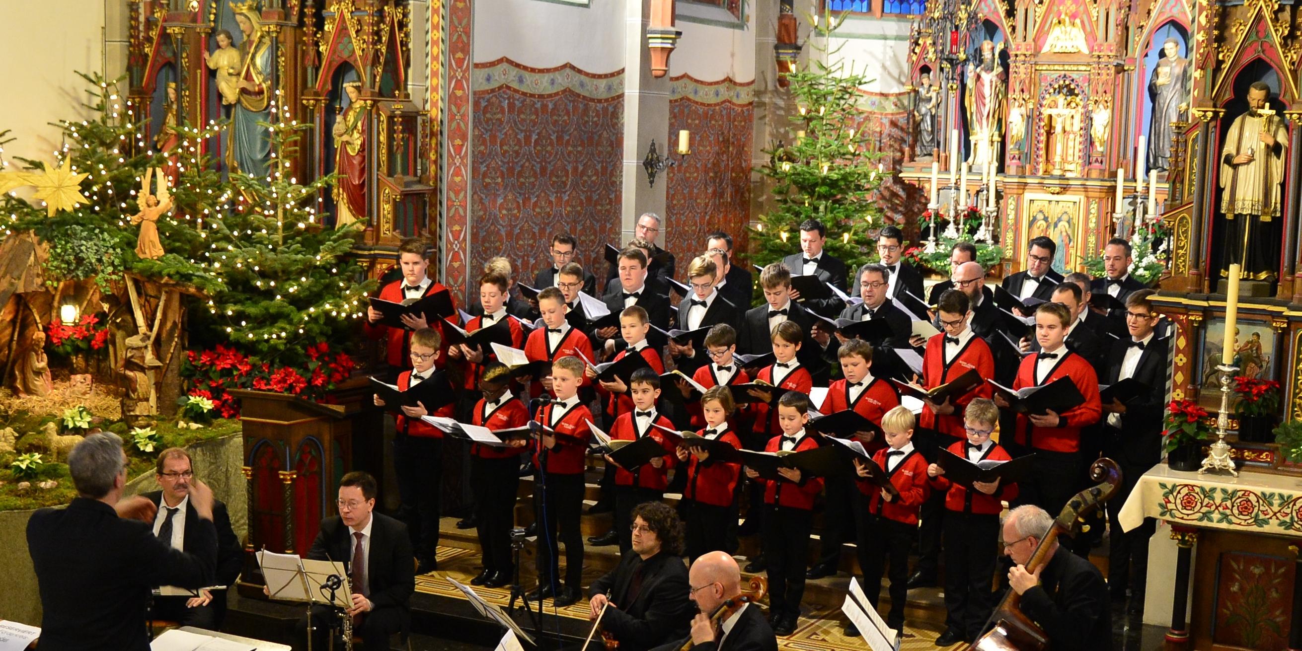 Viele Jungen und junge Männer stehen in einer Kirche und haben ihre Gesangsmappen in der Hand. Vor dem Chor spielt ein Orchester