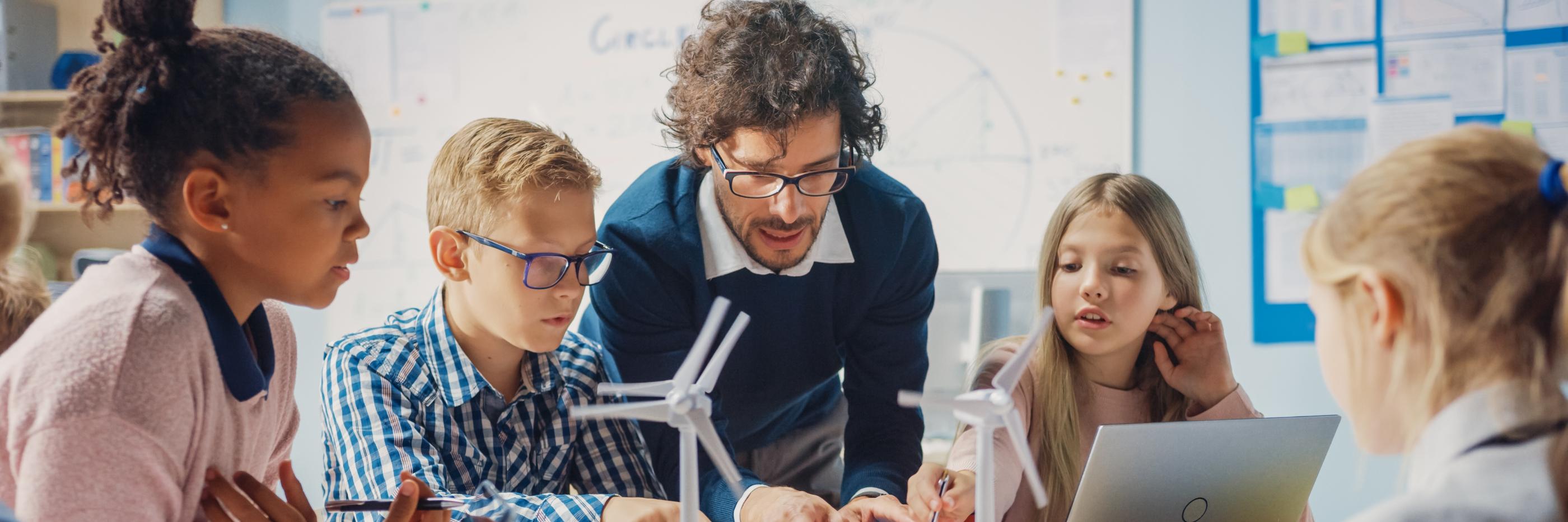 Man sieht Schüler*innen an einem Tisch mit Tablet, Laptop und Miniaturwindrädern. Ein Lehrer beugt sich zur Gruppe, um etwas zu erklären (c) shutterstock_1767368444 Man sieht Schüler*innen an einem Tisch mit Tablet, Laptop und Miniaturwindrädern. Ein Lehrer beugt sich zur Gruppe, um etwas zu erklären