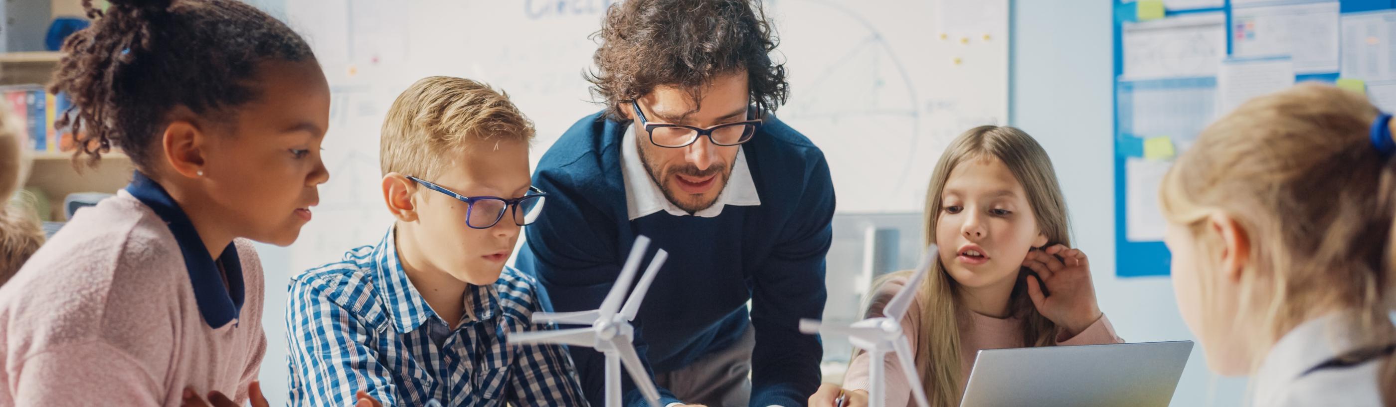 Man sieht Schüler*innen an einem Tisch mit Tablet, Laptop und Miniaturwindrädern. Ein Lehrer beugt sich zur Gruppe, um etwas zu erklären (c) shutterstock_1767368444 Man sieht Schüler*innen an einem Tisch mit Tablet, Laptop und Miniaturwindrädern. Ein Lehrer beugt sich zur Gruppe, um etwas zu erklären