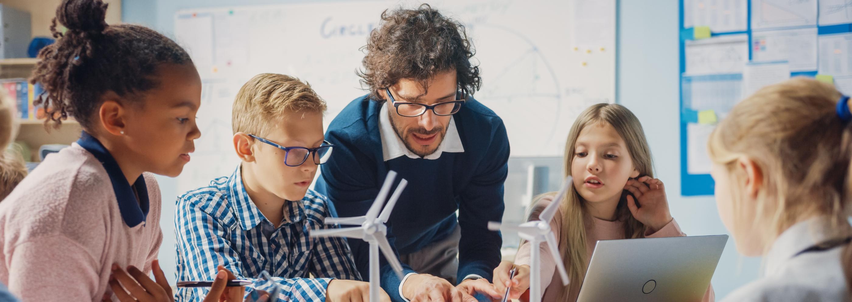 Man sieht Schüler*innen an einem Tisch mit Tablet, Laptop und Miniaturwindrädern. Ein Lehrer beugt sich zur Gruppe, um etwas zu erklären (c) shutterstock_1767368444 Man sieht Schüler*innen an einem Tisch mit Tablet, Laptop und Miniaturwindrädern. Ein Lehrer beugt sich zur Gruppe, um etwas zu erklären