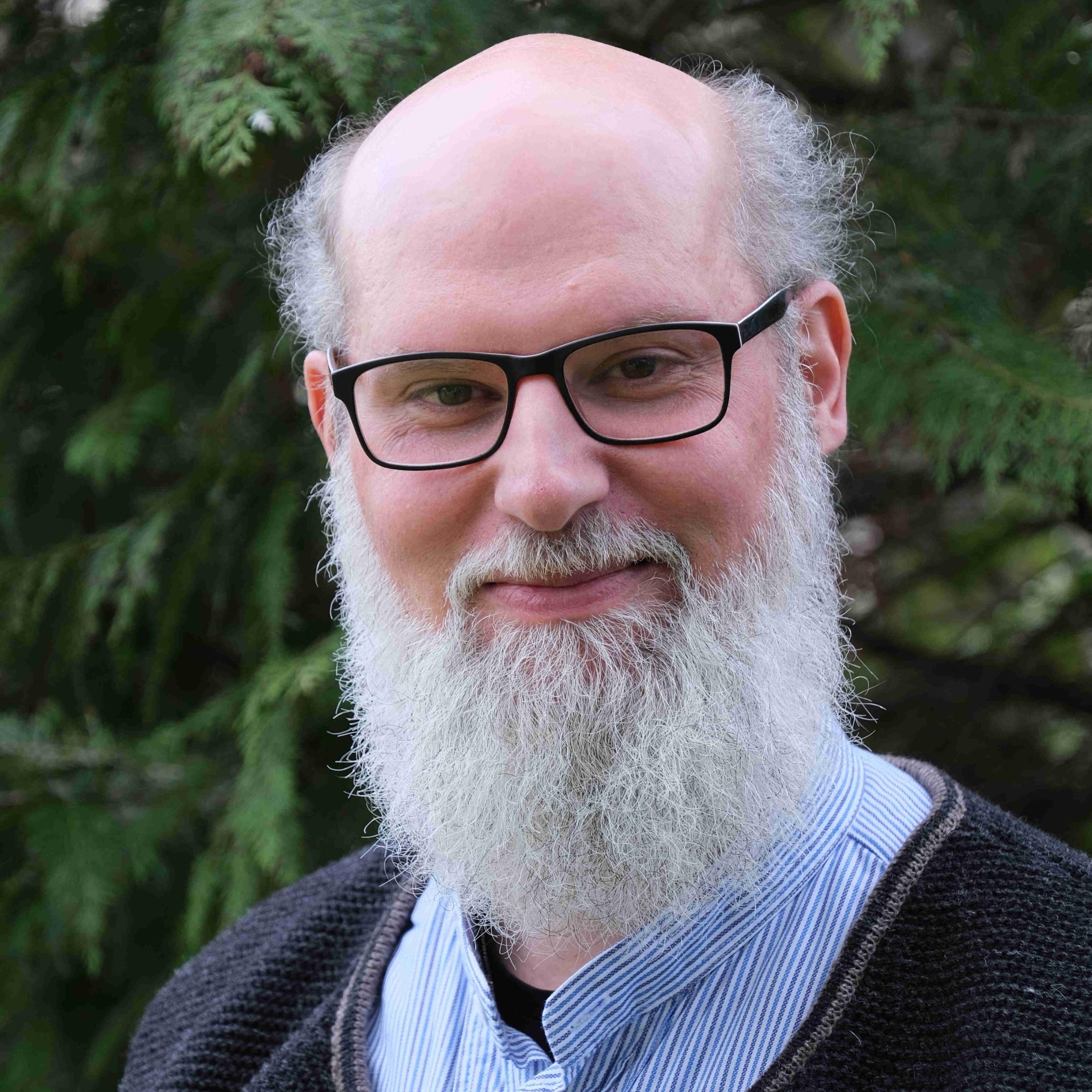 Ein Mann mit Brille und langem grauen Bart lächelt in die Kamera.
